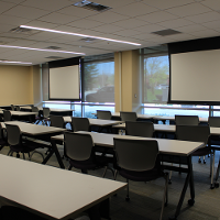 Tables and chairs are placed in a classroom setup facing two dropdown screens.