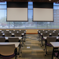 Tables and chairs are in a classroom setup in a large room facing two large projector screens.