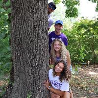 Four college students sitting next to and peeking out from behind a tree trunk.